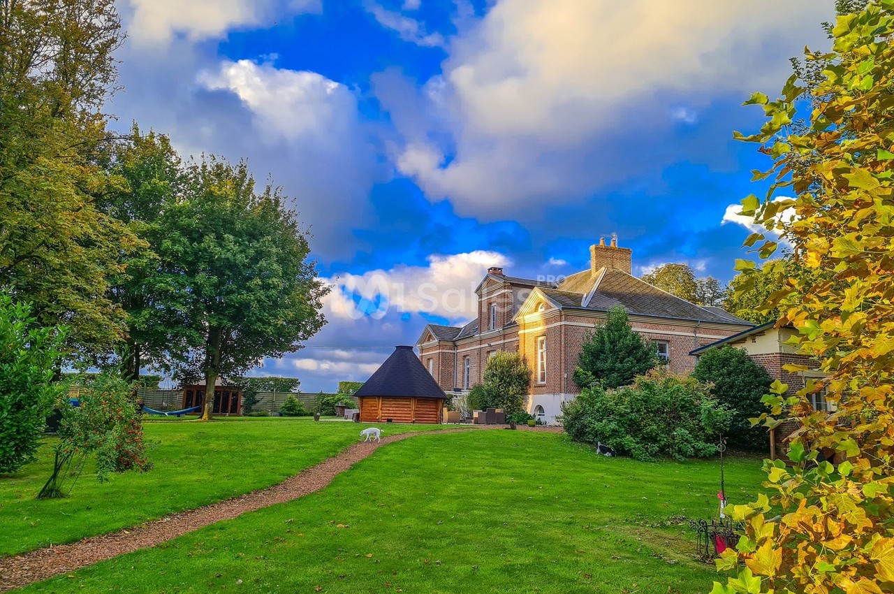 Grande maison en briques entourée d'un jardin verdoyant avec arbres, pelouse et ciel bleu parsemé de nuages.
