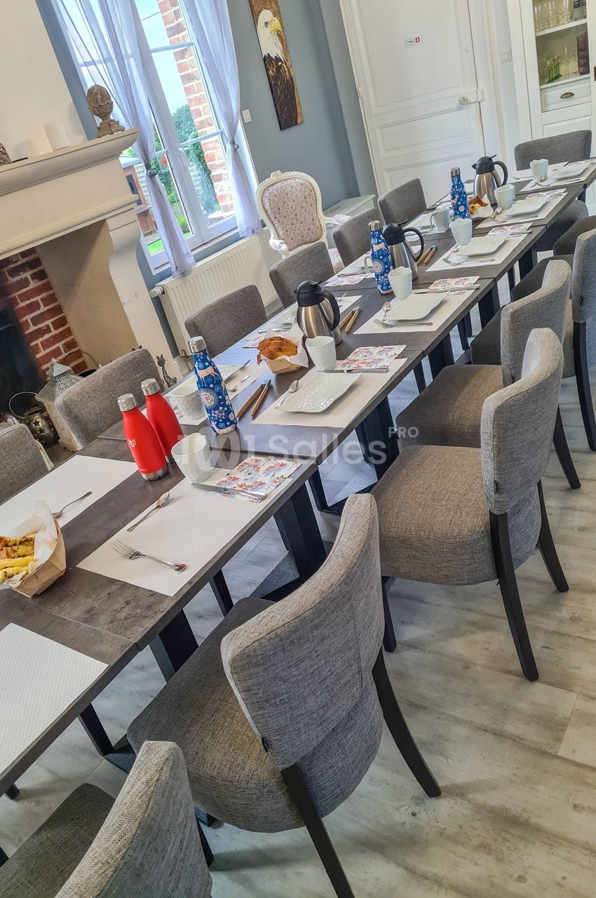 Table dressée pour un petit-déjeuner avec chaises grises, carafes, viennoiseries et vaisselle dans une salle lumineuse.