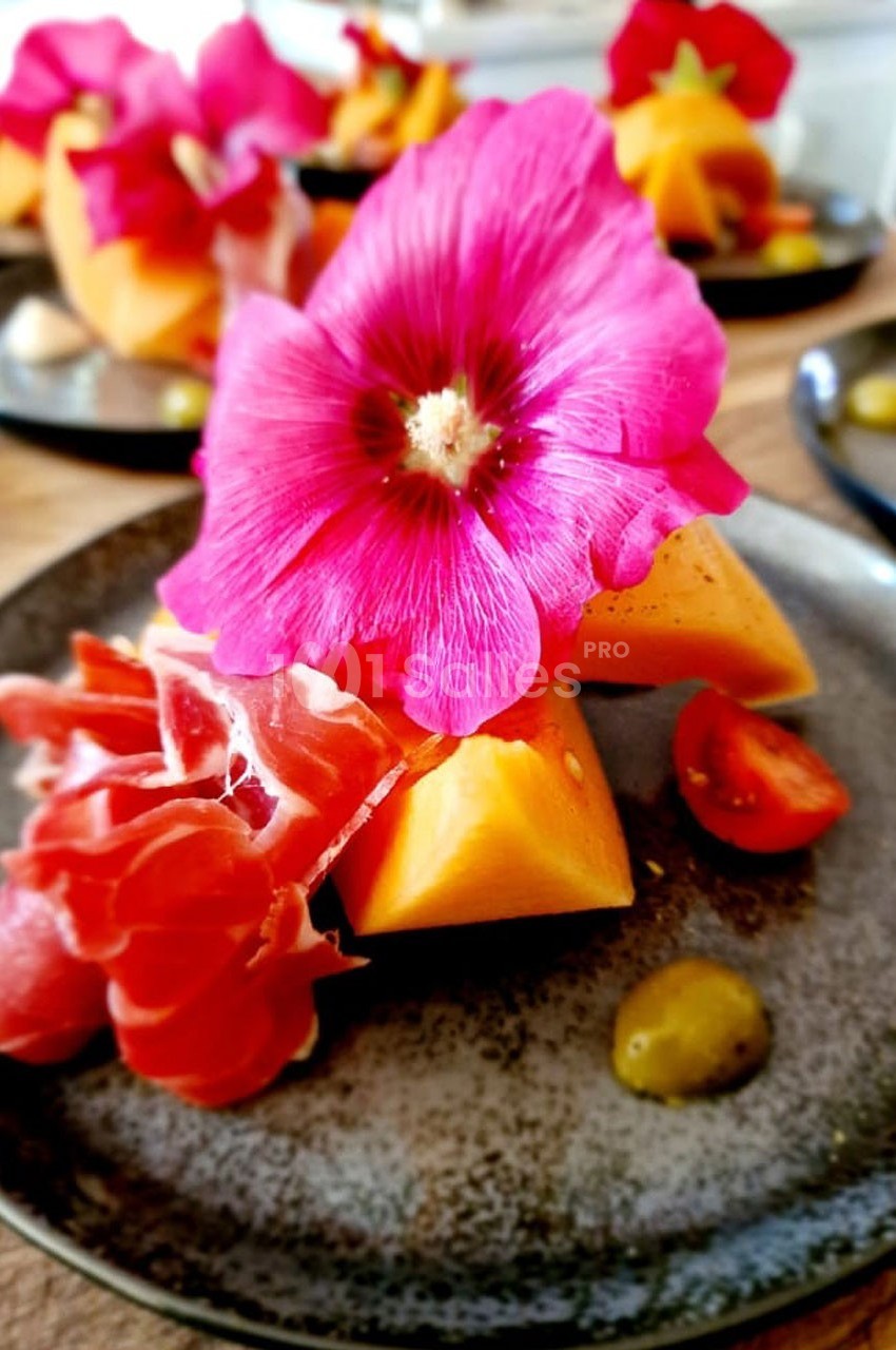 Assiette garnie de jambon cru, morceaux de melon, tomates cerises et une fleur comestible rose en décoration.