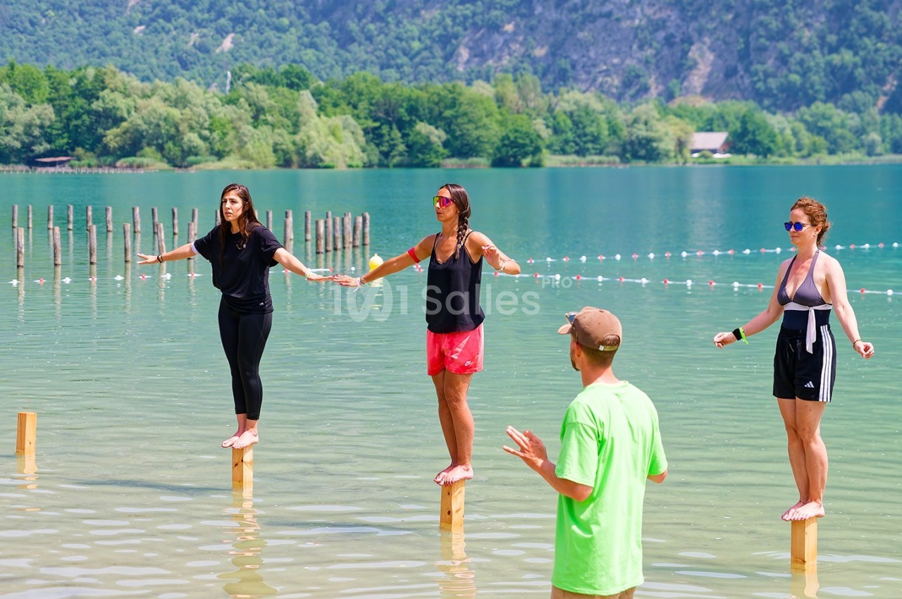 Trois femmes debout sur des poteaux dans un lac peu profond, encadrées par un homme en t-shirt vert, avec un paysage…