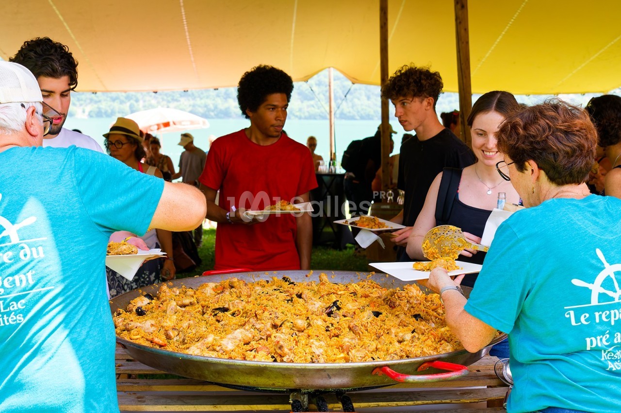 Des personnes se servent de la paella dans un grand plat lors d'un événement en plein air sous une tente.