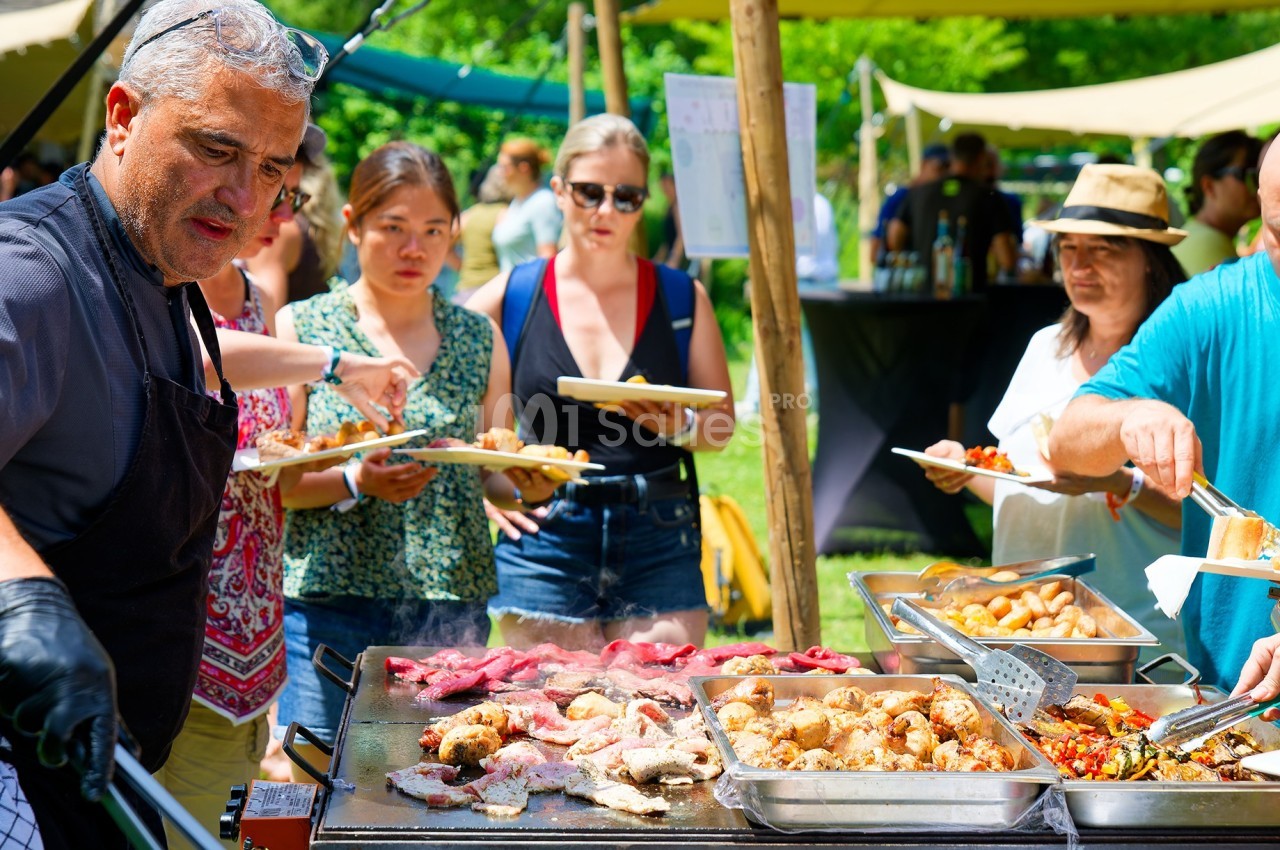 Un cuisinier prépare de la viande et des légumes sur une plancha lors d'un événement en plein air avec des participants.