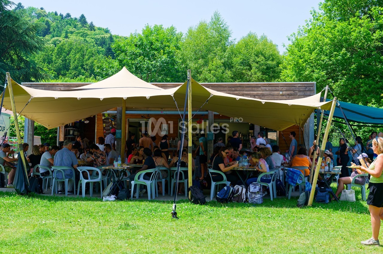 Un groupe de personnes assises et debout sous une grande tente beige dans un espace vert en plein air.