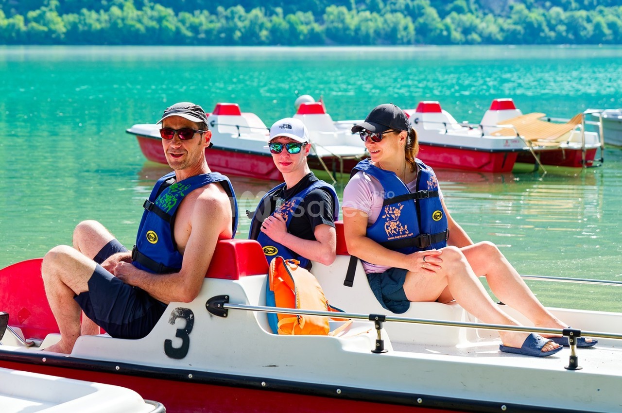 Trois personnes assises sur un pédalo au bord d'un lac turquoise, portant des gilets de sauvetage.