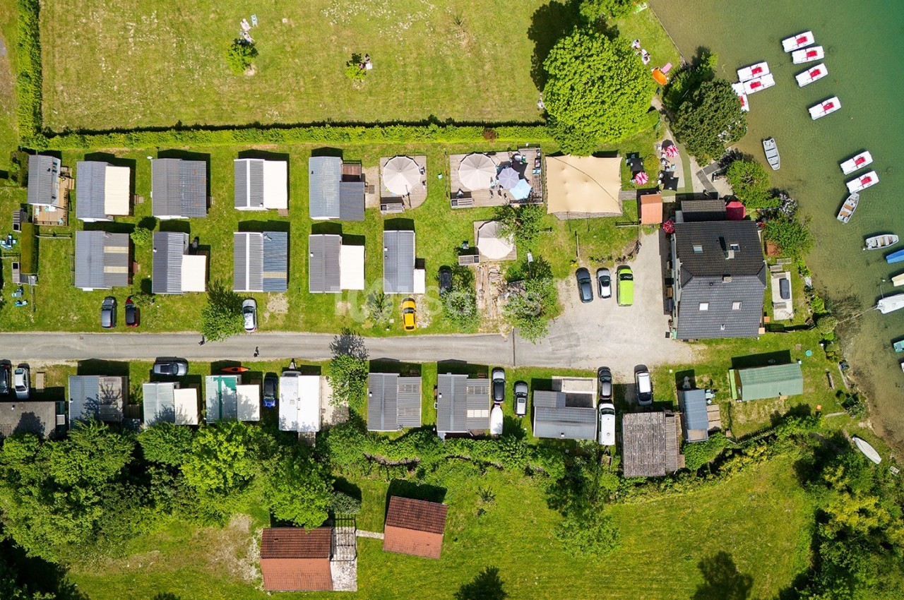 Vue aérienne d'un camping avec emplacements, bâtiments, voitures et accès direct à un lac avec des bateaux amarrés.