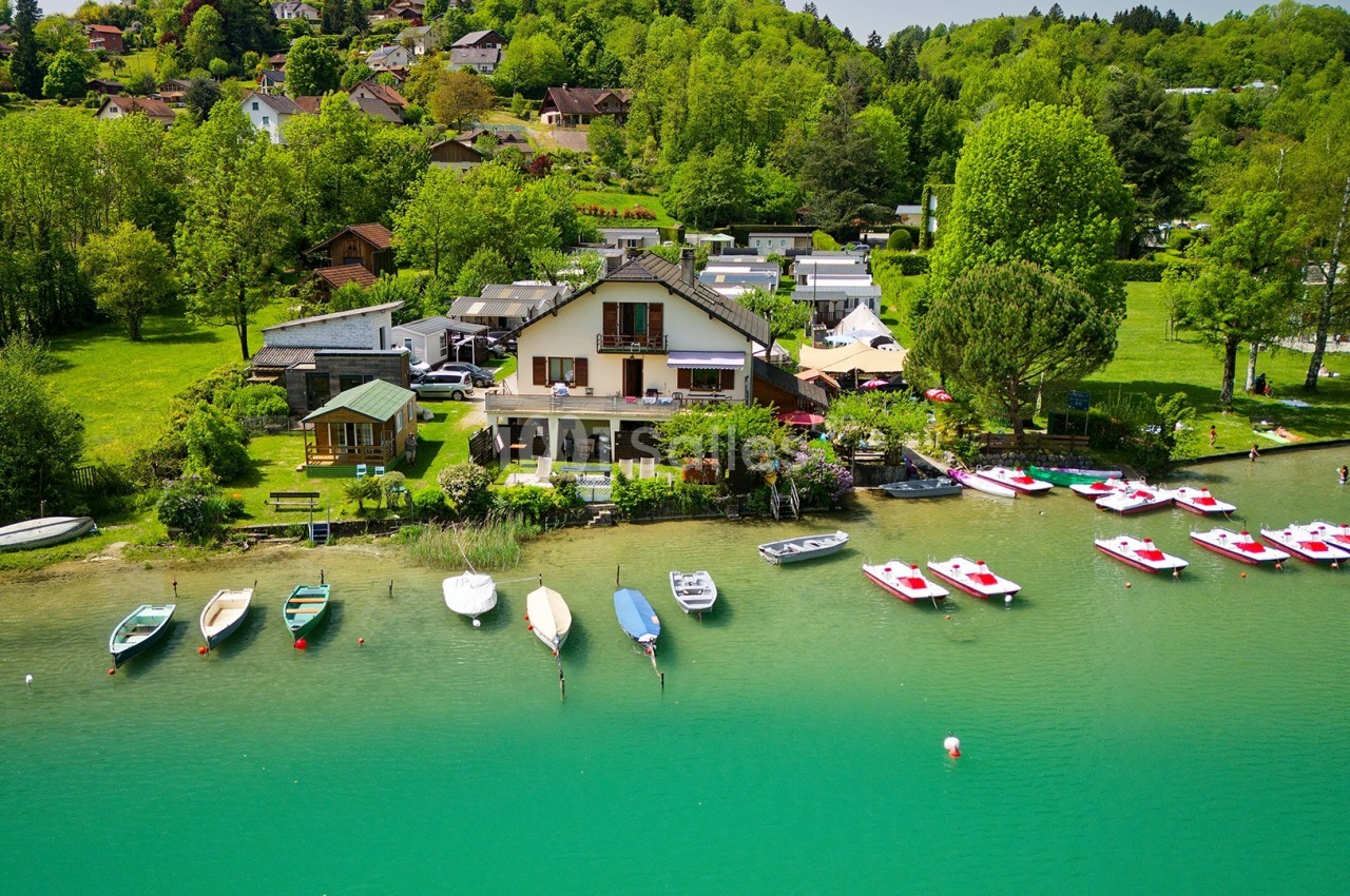 Vue d'une maison au bord d'un lac avec des bateaux amarrés et une végétation dense en arrière-plan.