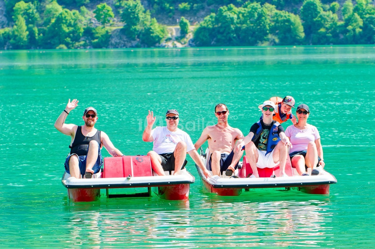 Un groupe de personnes souriantes sur deux pédalos rouges flottant sur un lac turquoise entouré de végétation.