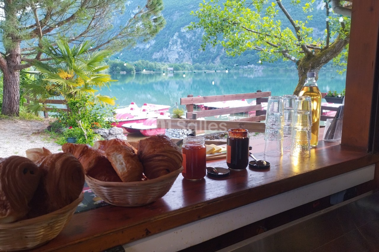 Vue depuis un comptoir avec viennoiseries, confitures et verres, donnant sur un lac entouré de montagnes et de verdure.