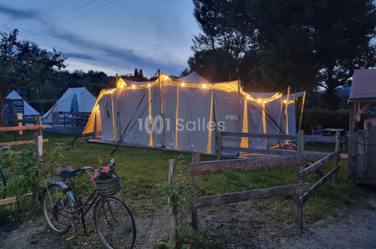 Tente éclairée au crépuscule dans un camping, avec une clôture en bois et un vélo garni d'un panier fleuri au premier plan.