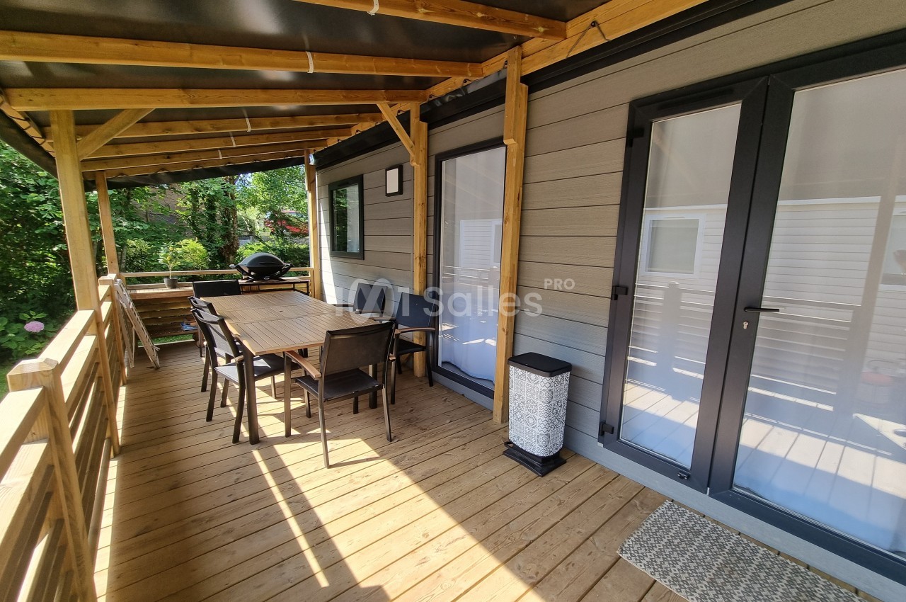 Terrasse en bois couverte avec table, chaises, barbecue et accès à une habitation par des portes vitrées.