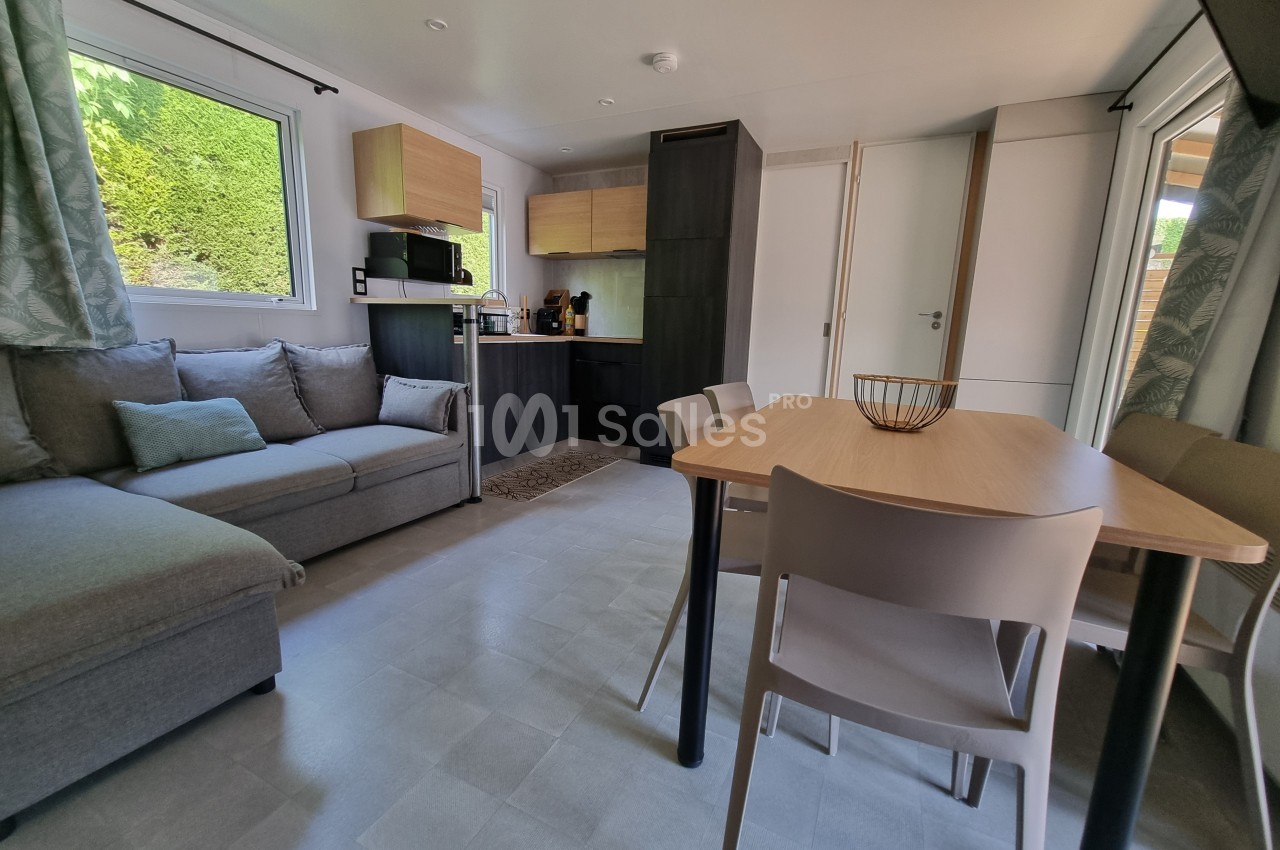 Intérieur lumineux d'un mobil-home avec salon, table à manger, cuisine équipée et grandes fenêtres donnant sur l'extérieur.