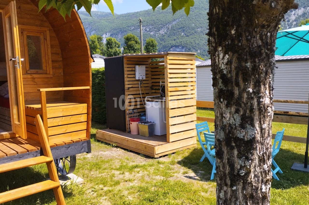 Cabane en bois avec terrasse, petite structure technique à côté, entourées de verdure et montagnes en arrière-plan.