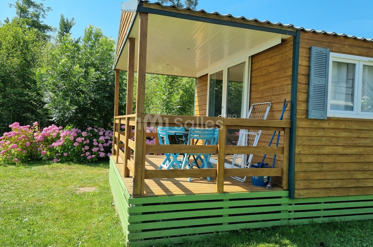 Mobil-home en bois avec terrasse couverte, table et chaises bleues, entouré de verdure et de fleurs roses.