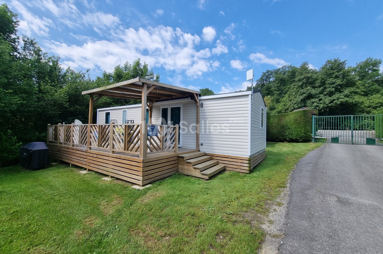 Mobil-home blanc avec terrasse en bois, situé sur une pelouse près d'une route et entouré de verdure.