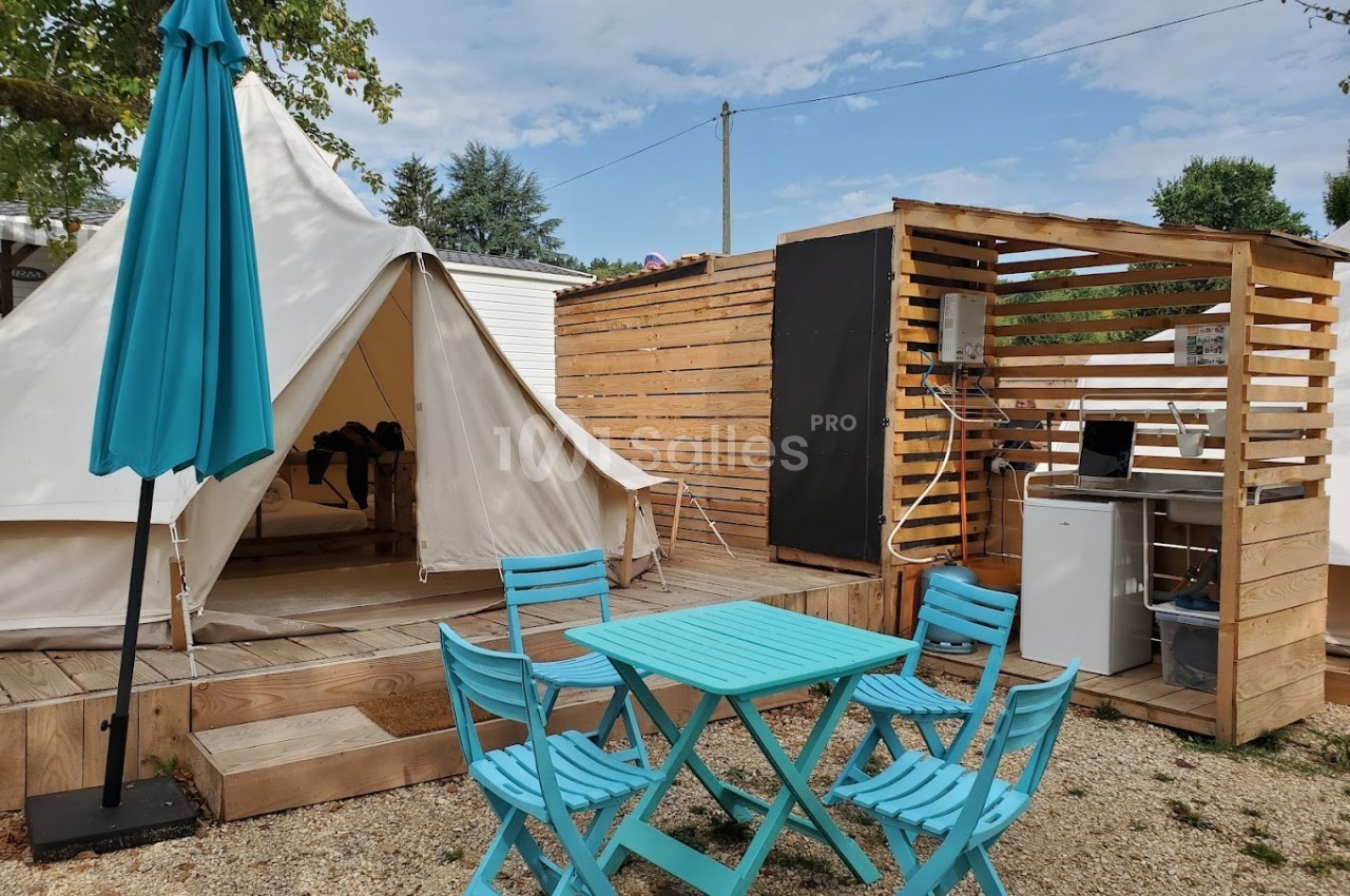 Tente en toile avec terrasse en bois, mobilier de jardin bleu et espace cuisine extérieur dans un cadre ensoleillé.