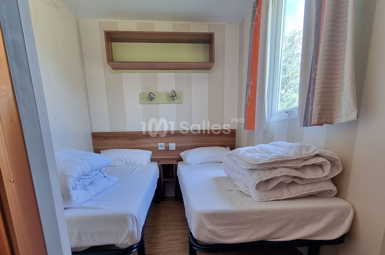 Chambre lumineuse avec deux lits simples, draps pliés, étagère murale et fenêtre donnant sur l'extérieur.