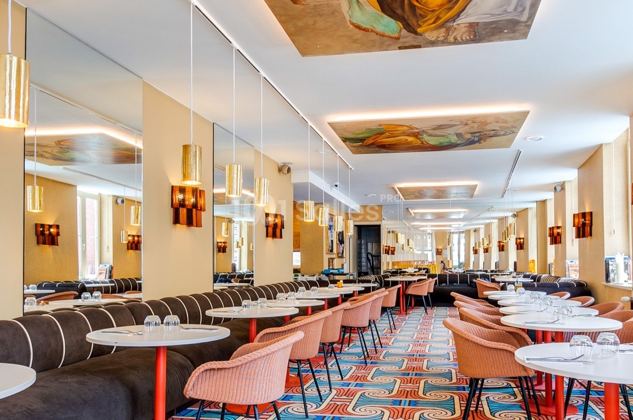 Salle de restaurant lumineuse avec tables rondes, chaises colorées, fresques au plafond et décorations modernes.