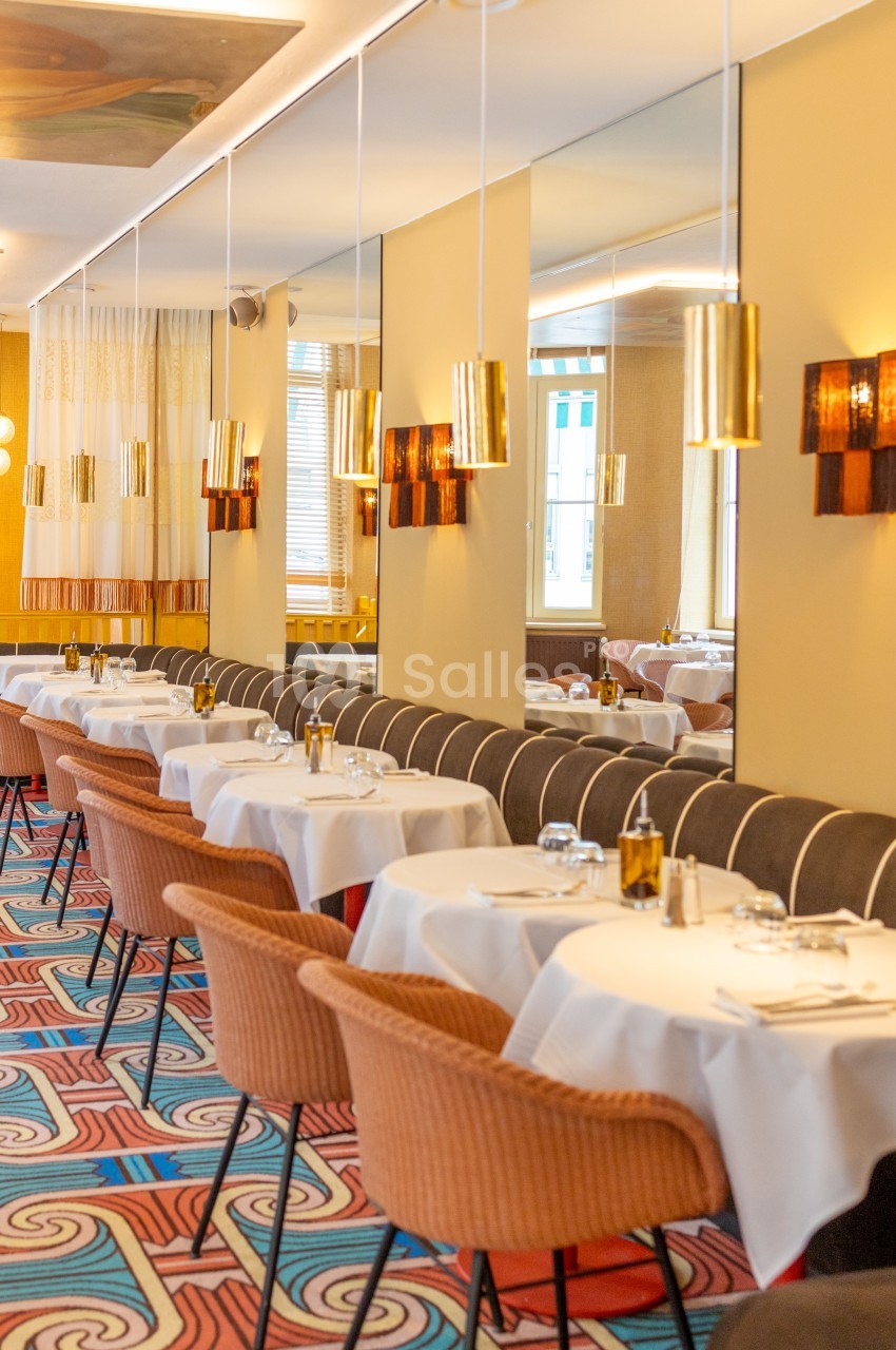 Salle de restaurant élégante avec tables dressées, chaises en velours, miroirs muraux et luminaires suspendus.