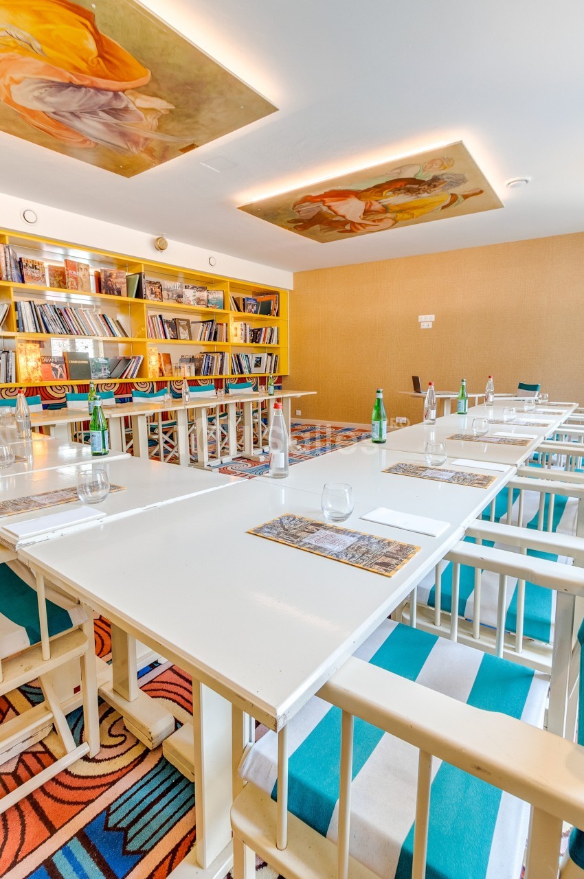 Salle de réunion lumineuse avec tables blanches, chaises colorées, étagères remplies de livres et fresques au plafond.