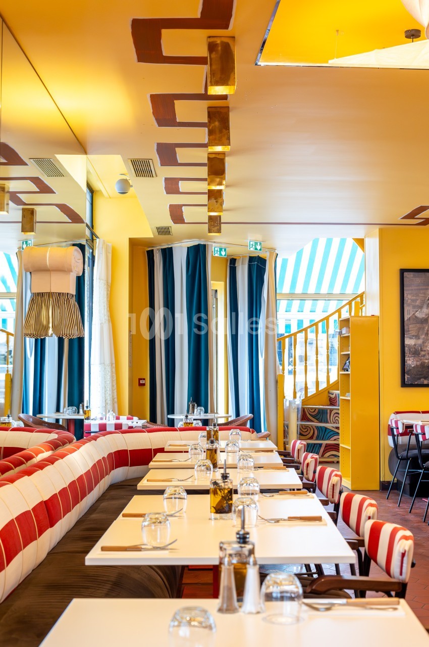 Salle de restaurant lumineuse avec tables alignées, chaises rayées, murs jaunes et escalier en colimaçon au fond.