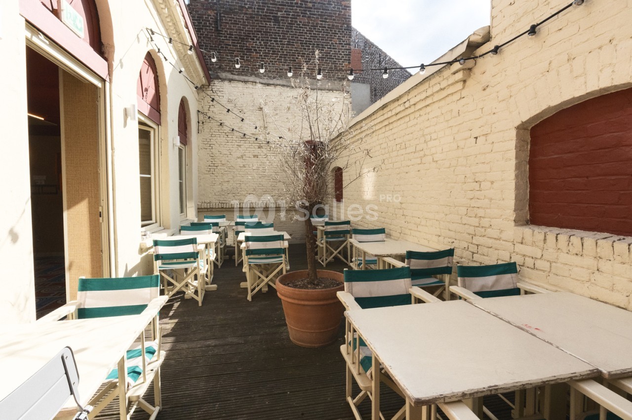 Cour intérieure avec tables blanches et chaises à dossier vert, murs de briques clairs, guirlande lumineuse et jardinière.