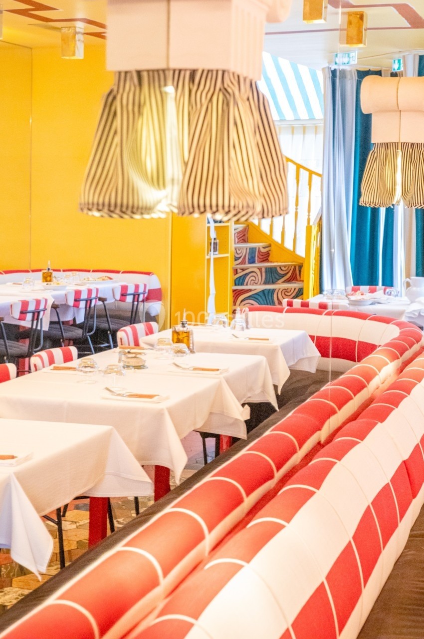 Salle de restaurant lumineuse avec banquettes rouges et blanches, tables dressées et décor coloré.
