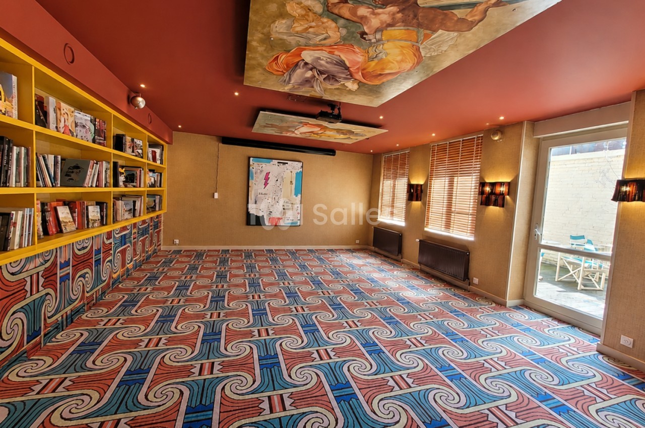 Salle intérieure aux murs ocre et plafond rouge ornementé, bibliothèque jaune, tapis géométrique bleu-rouge.