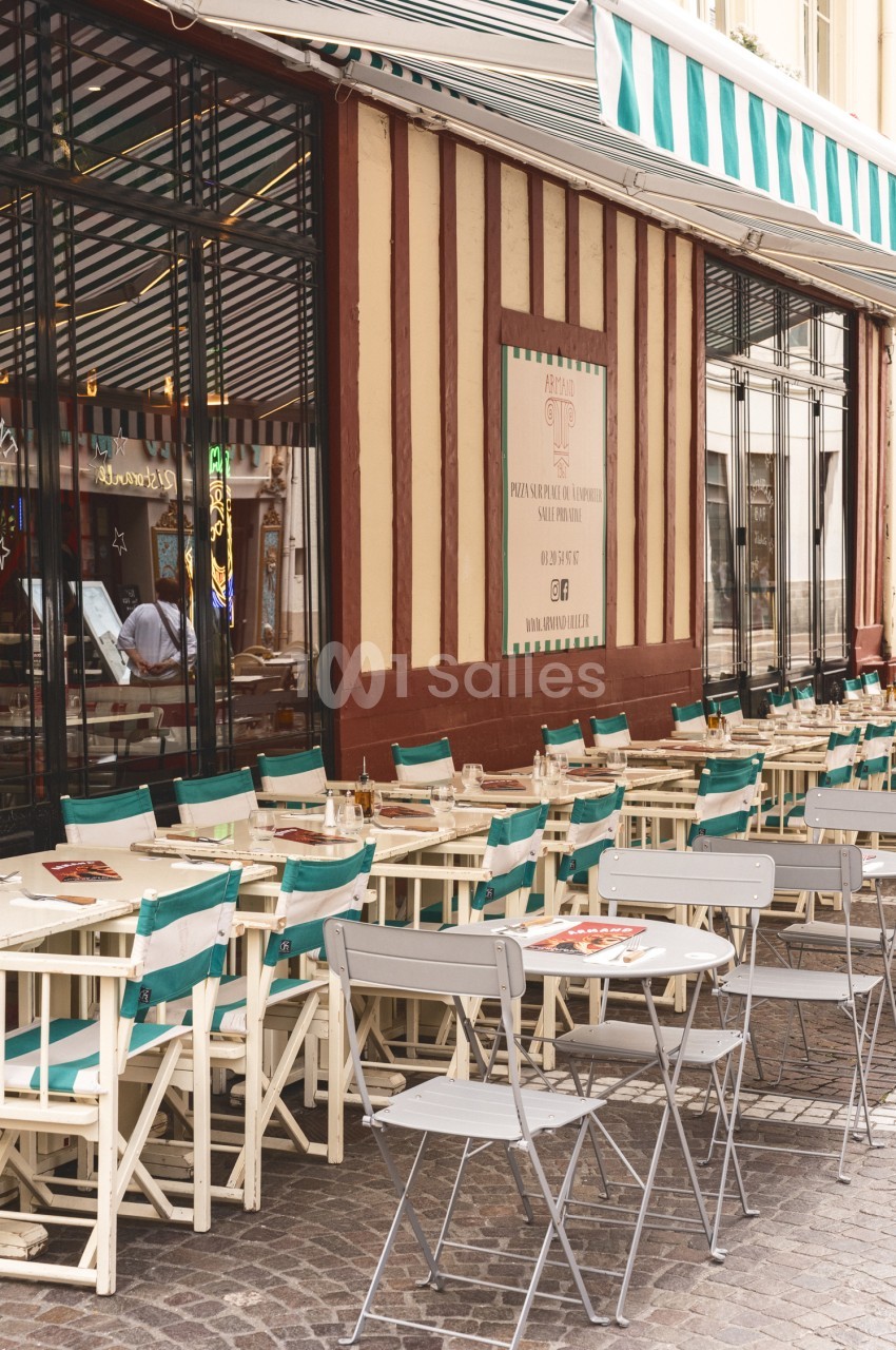 Terrasse extérieure avec tables dressées, chaises turquoise et blanches, façade à colombages beige et rouge.