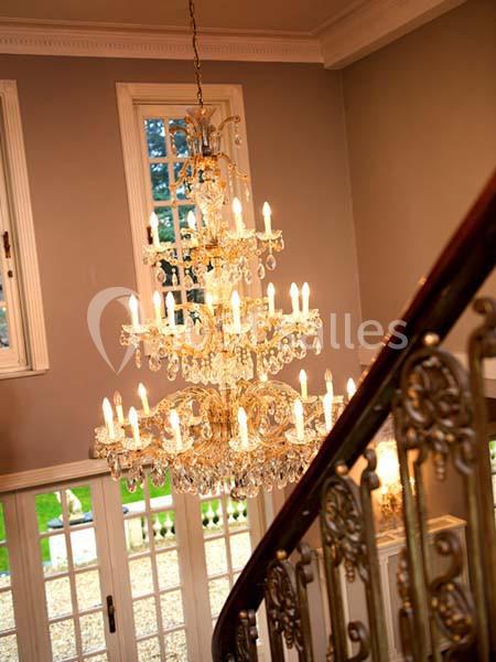 Lustre en cristal allumé suspendu dans une entrée avec escalier en bois et rambarde ornée de motifs dorés.