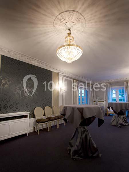 Salle élégante avec tables hautes drapées, chaises alignées contre un mur décoré, lustre central et fenêtres lumineuses.