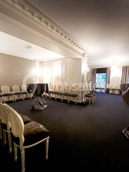 Salle élégante avec chaises alignées le long des murs, tables hautes couvertes de nappes et lumière naturelle en arrière…