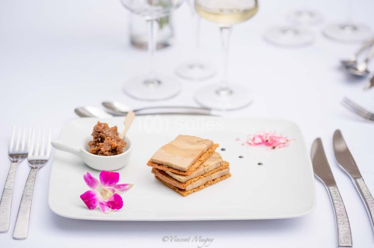 Assiette gastronomique avec foie gras sur pain d'épices, chutney, fleur décorative et verres en arrière-plan.