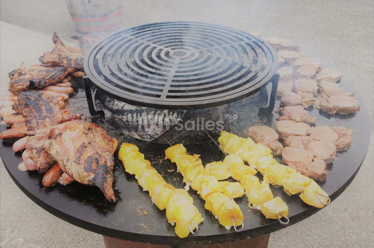 Grill circulaire avec diverses viandes en cuisson, dont brochettes de poulet, saucisses et côtes.