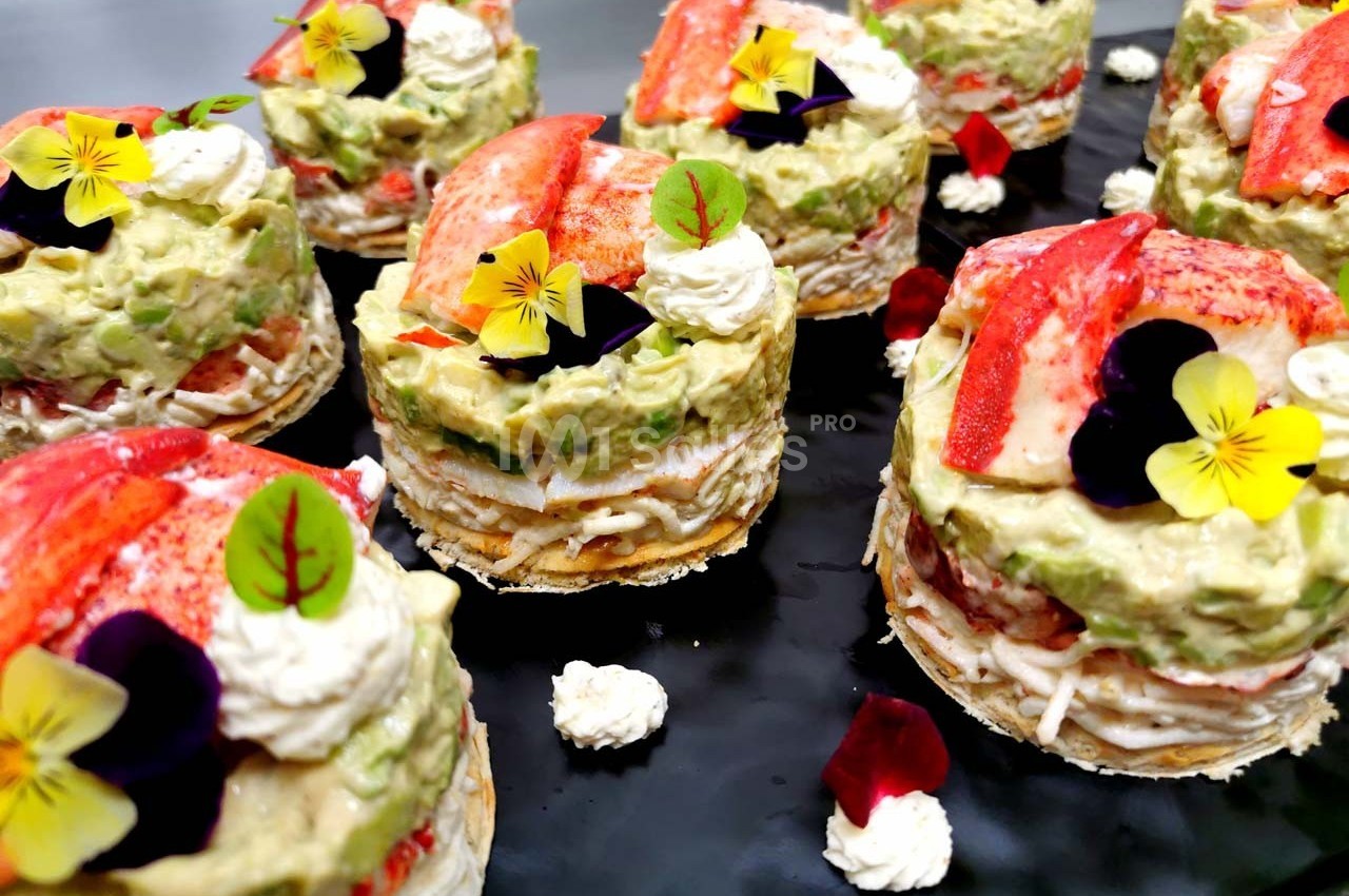 Assortiment de millefeuilles salés garnis de crème, avocat, morceaux de homard et décorés de fleurs comestibles.