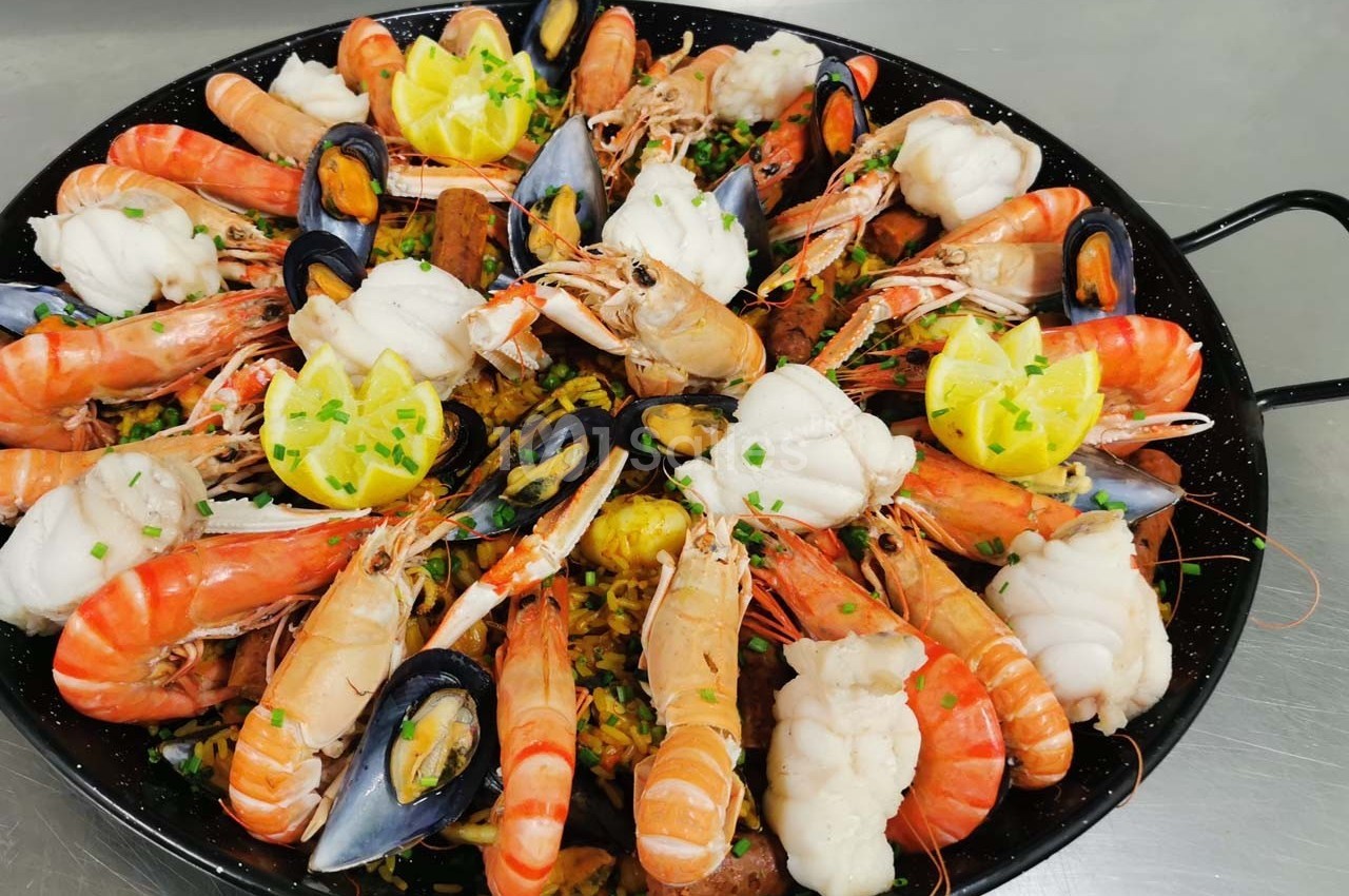 Plat de paella garni de crevettes, moules, morceaux de poisson et quartiers de citron, présenté dans une poêle.