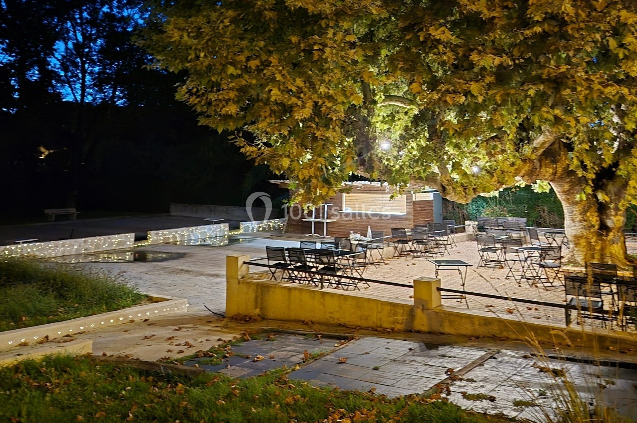 Terrasse éclairée en soirée avec des tables et chaises disposées sous un grand arbre, près d'un comptoir en bois.