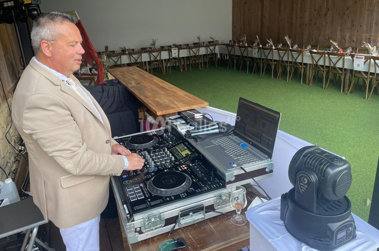 Un DJ mixe de la musique sur une table de mixage dans un espace extérieur avec des tables décorées en arrière-plan.