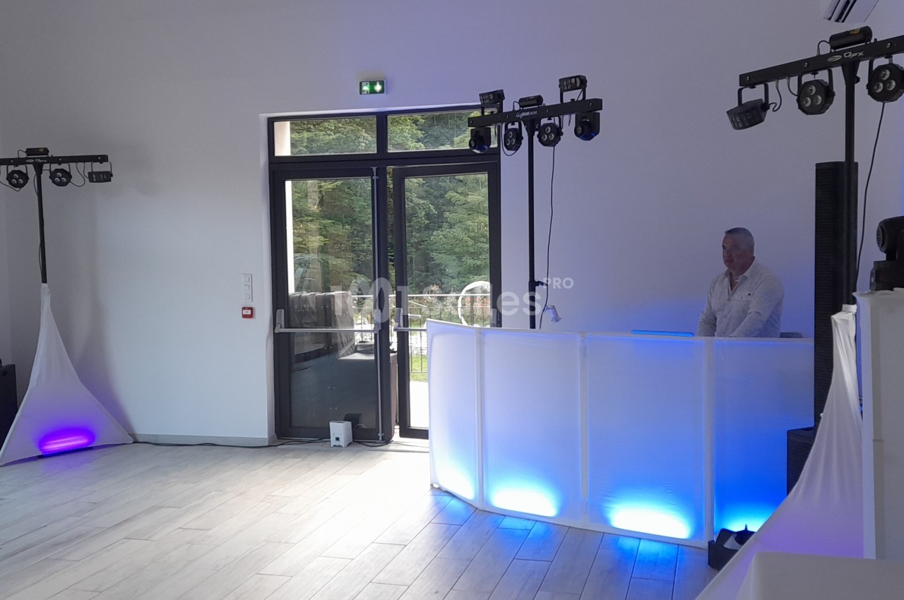 Un DJ derrière une table lumineuse blanche dans une salle équipée de lumières et d'enceintes pour un événement.