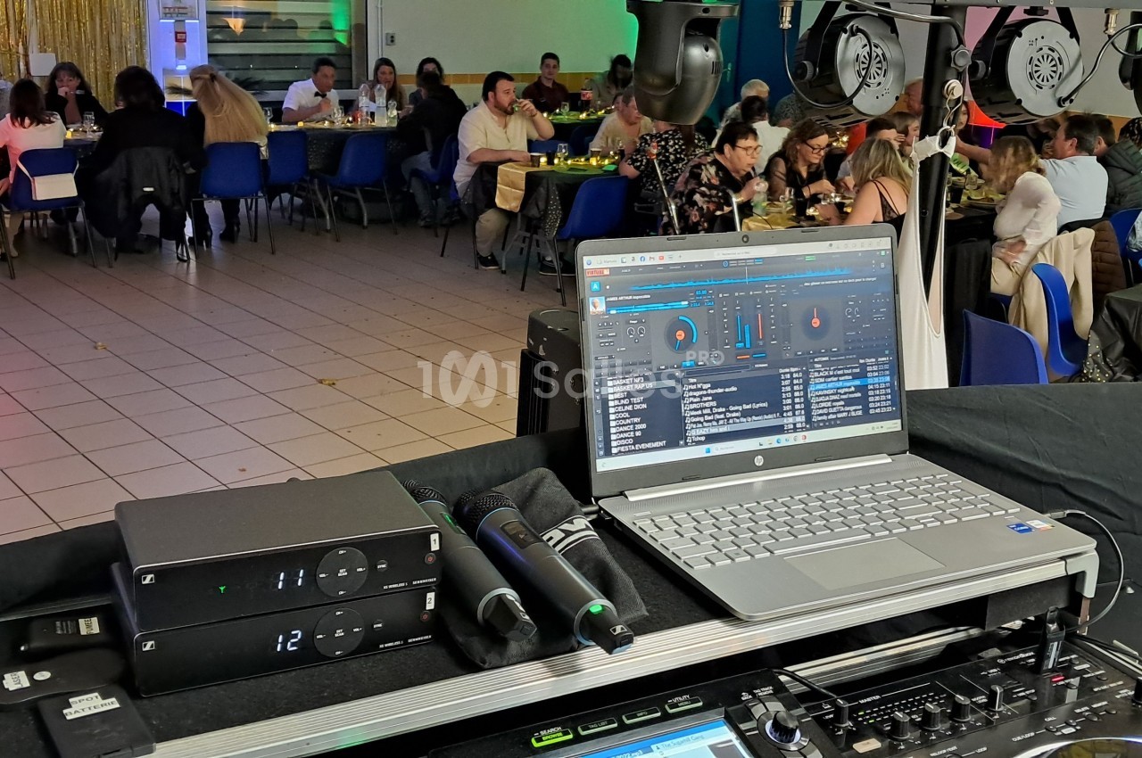 Vue d'une salle de réception avec des invités attablés et un poste de DJ en premier plan, équipé d'un ordinateur portable.