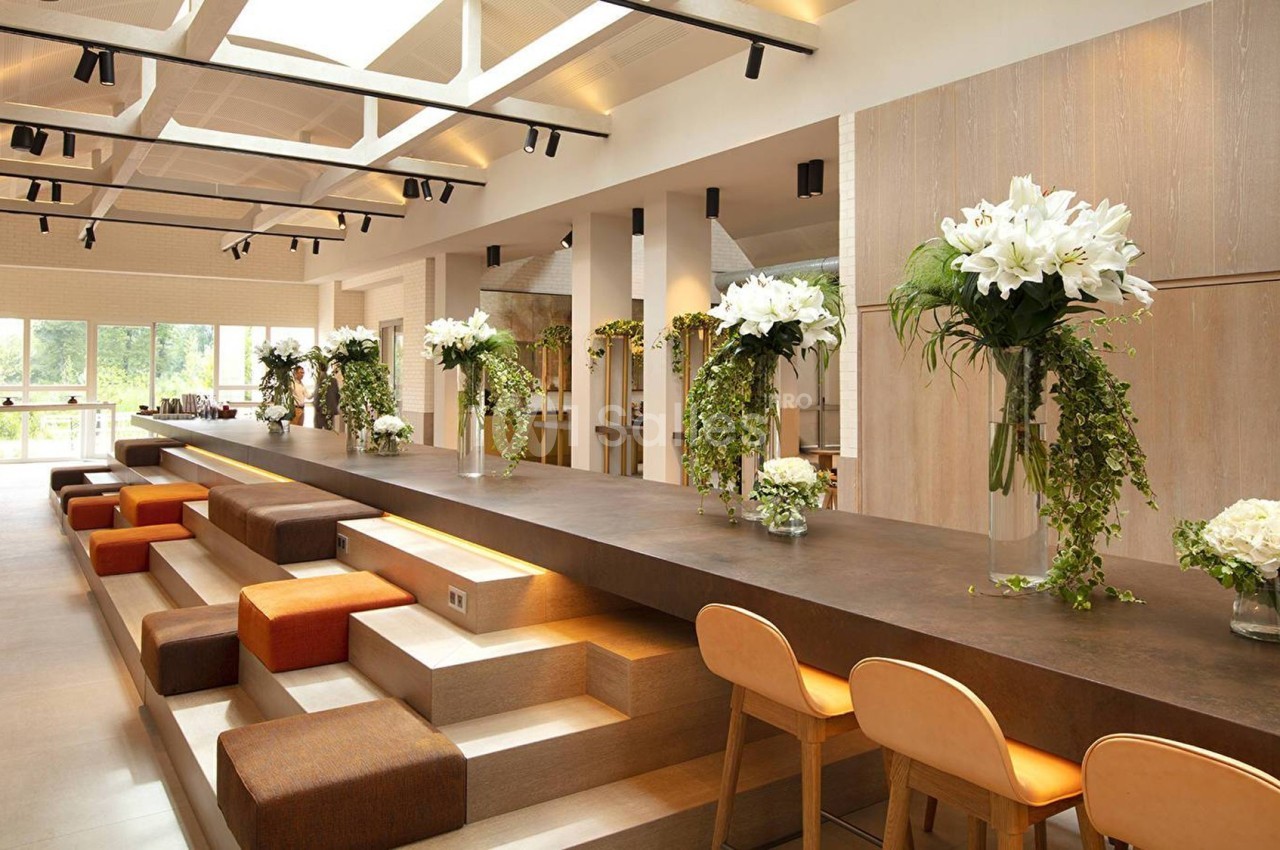 Salle lumineuse avec un long comptoir en bois, décorée de fleurs blanches et vertes, entourée de chaises et banquettes.