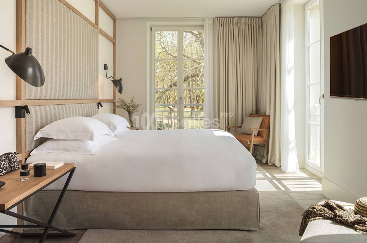 Chambre lumineuse avec lit double, bureau en bois, fauteuil, grande fenêtre et rideaux beiges laissant entrer la lumière.