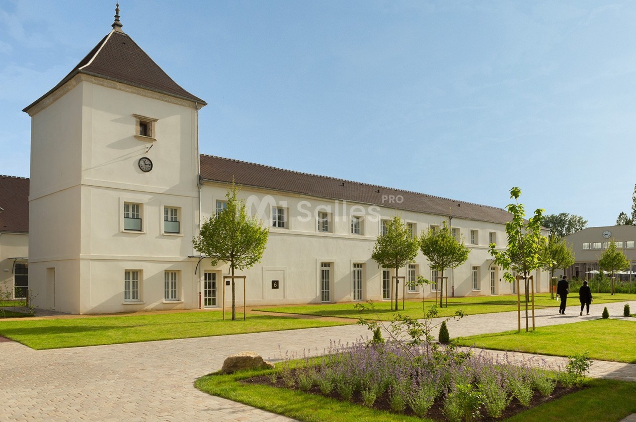 Bâtiment blanc à deux étages avec une tour, entouré d'espaces verts et de chemins pavés, sous un ciel dégagé.