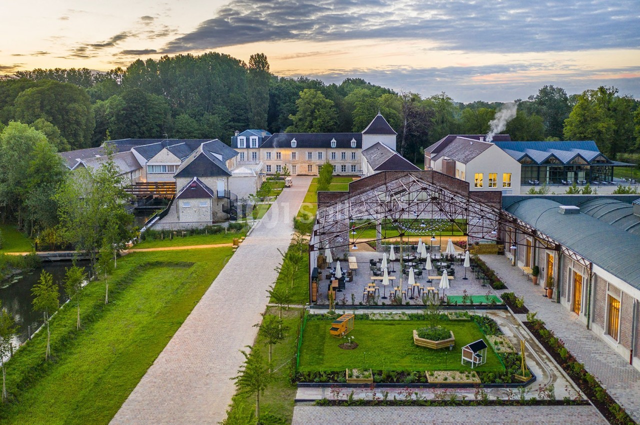 Vue aérienne d'un domaine avec bâtiments modernes et anciens entourés de verdure, sous un ciel partiellement nuageux.
