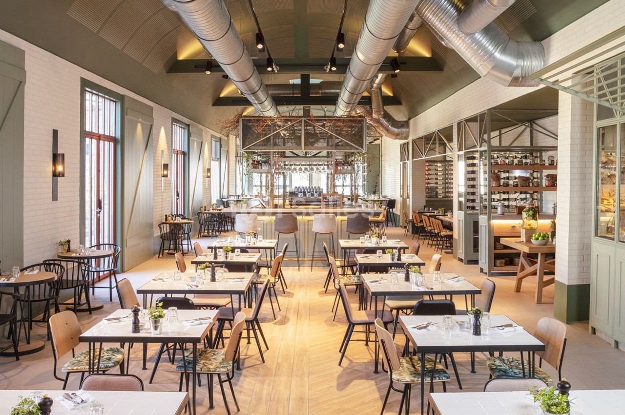 Salle de restaurant lumineuse avec tables dressées, chaises variées, plafond industriel et cuisine ouverte en arrière-plan.