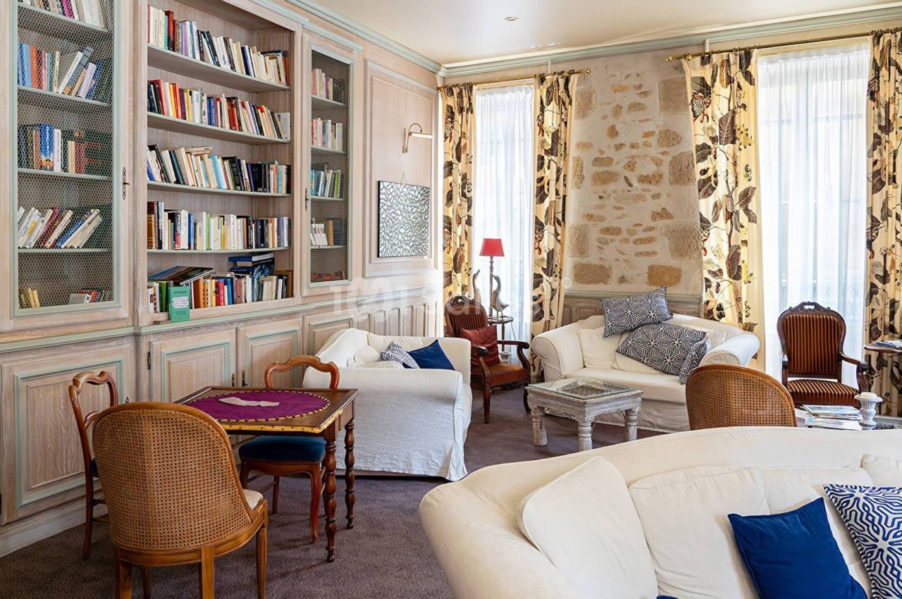 Salon lumineux avec bibliothèque en bois, fauteuils et canapés, murs en pierre et grandes fenêtres avec rideaux fleuris.