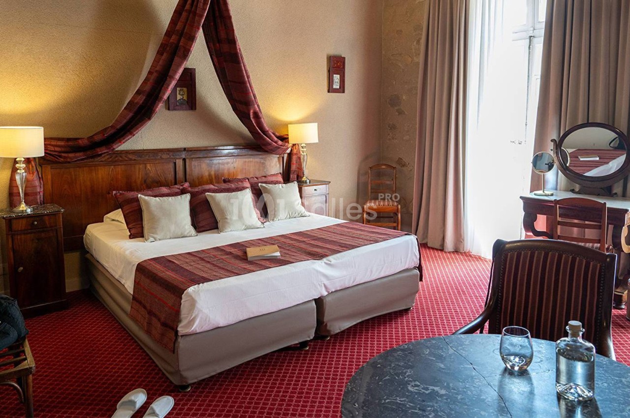 Chambre d'hôtel avec lit double, décorée de tons rouges, équipée de lampes, d'une coiffeuse et d'une table avec carafe d'eau.