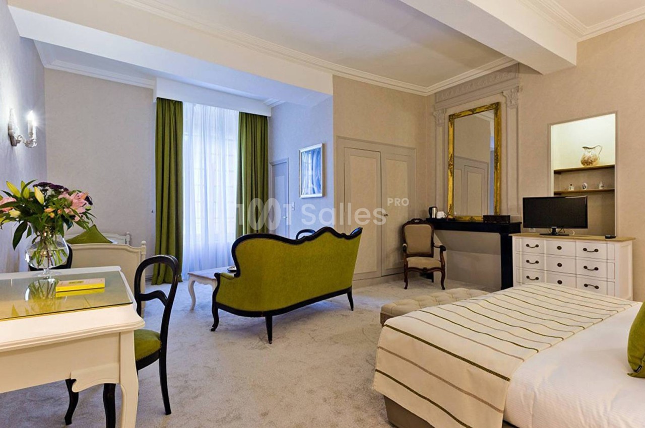 Chambre lumineuse avec lit double, canapé vert, bureau, télévision et grande fenêtre avec rideaux verts.