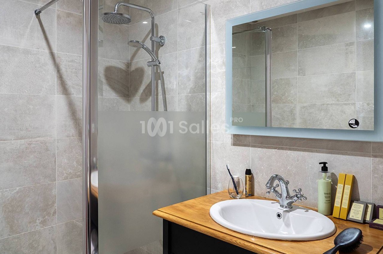Salle de bain avec douche à paroi vitrée, lavabo sur meuble en bois, miroir éclairé et accessoires de toilette.