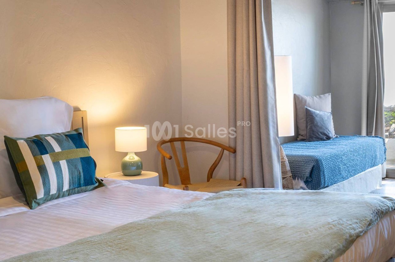 Chambre lumineuse avec lit double, lit d'appoint, fauteuil en bois, lampe de chevet et accès à une terrasse.