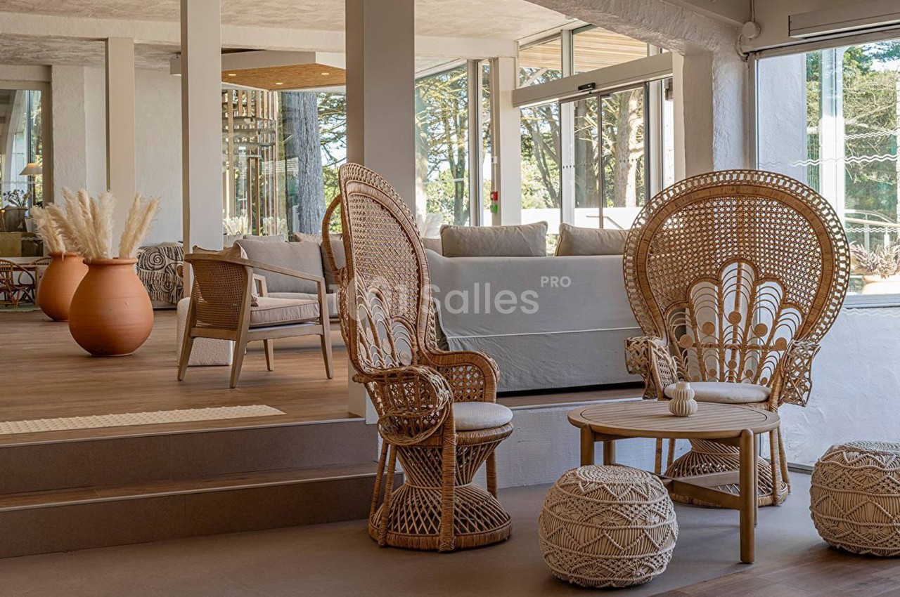 Espace intérieur lumineux avec fauteuils en rotin, table basse, poufs tressés et grande jarre décorative en arrière-plan.